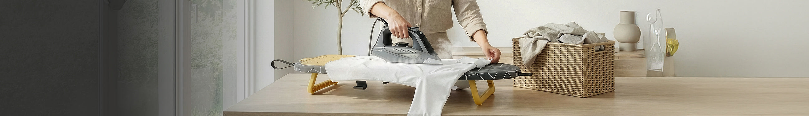Pessoa a passar roupa numa tábua de engomar de mesa, compacta e dobrável da marca Flama com ferro a vapor ao lado de um cesto de roupa em vime